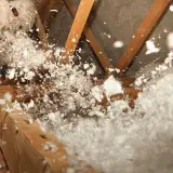 Un ouvrier en combinaison souffle de la laine de verre dans un grenier, créant un nuage blanc et cotonneux pour une isolation efficace.