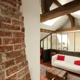 Murs en briques apparentes et poutres de bois, ambiance chaleureuse pour cette rénovation combles. Canapé blanc, coussins rouges, style loft.