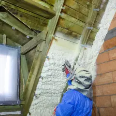 Un ouvrier en combinaison bleue applique du polyuréthane projeté sur la charpente d'un grenier, isolant ainsi les murs en briques et le toit.