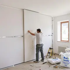 Pose d'une cloison intérieure pour un meilleur isolation phonique. Un ouvrier ajuste un panneau blanc sur une structure métallique.