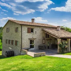 Belle ferme en pierre, rénovation réussie, avec un porche couvert et des roses épanouies. Un paysage verdoyant et un ciel bleu complètent ce tableau.