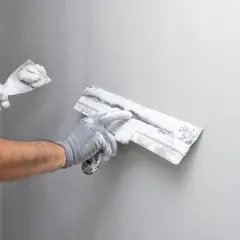 Mains gantées appliquant du machefer sur un mur gris avec des truelles.