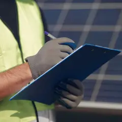 Un technicien en 50549, portant un gilet de sécurité, note des informations sur un presse-papiers bleu devant des panneaux solaires.