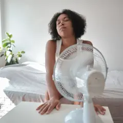 Une jeune femme en débardeur blanc, accablée par la chaleur de l'été, cherche un peu de fraîcheur devant un ventilateur. L'isolation de sa chambre ne suffit pas.