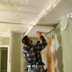 Un homme installe un panneau au plafond, entouré de bâches. Attention, la laine de verre peut présenter un danger pour les poumons si mal manipulée.
