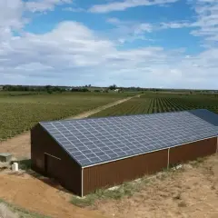 Vue aérienne d'un hangar brun avec un toit couvert de panneaux photovoltaïques, entouré de vignobles.