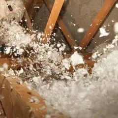 Un ouvrier en combinaison souffle de la laine de verre dans un grenier, créant un nuage blanc et cotonneux pour une isolation efficace.