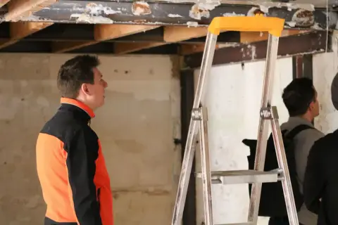 Un homme en veste orange et noire observe le plafond délabré d'un bâtiment, signe d'une future rénovation énergétique des bâtiments publics.