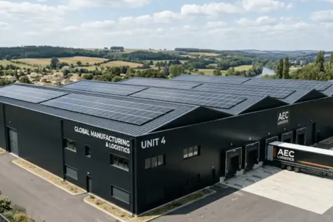 Bâtiment industriel noir avec panneaux solaires sur le toit, portant l'inscription "GLOBAL MANUFACTURING & LOGISTICS UNIT 4". Un camion AEC LOGISTICS est garé à quai.