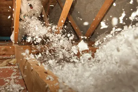 Un ouvrier en combinaison souffle de la laine de verre dans un grenier, créant un nuage blanc et cotonneux pour une isolation efficace.