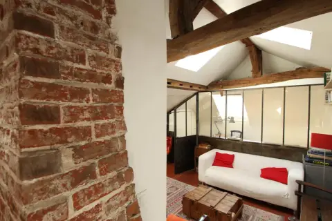 Murs en briques apparentes et poutres de bois, ambiance chaleureuse pour cette rénovation combles. Canapé blanc, coussins rouges, style loft.