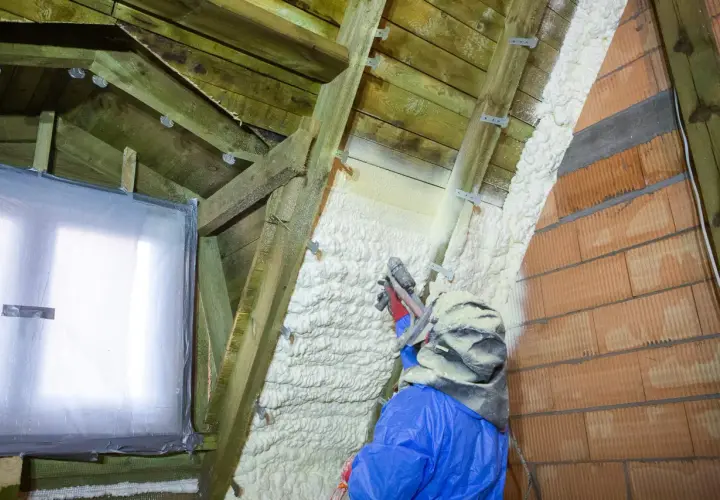 Un ouvrier en combinaison bleue applique du polyuréthane projeté sur la charpente d'un grenier, isolant ainsi les murs en briques et le toit.