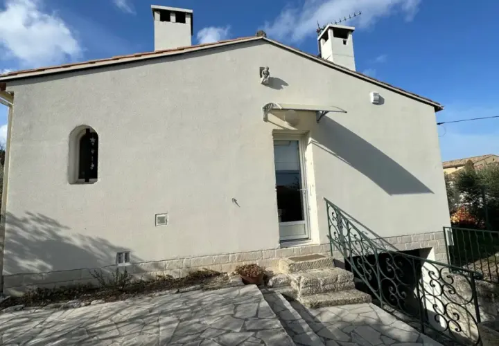 Maison Phénix 1970, blanche, avec cheminées, sous un ciel bleu. Escalier en pierre menant à la porte.