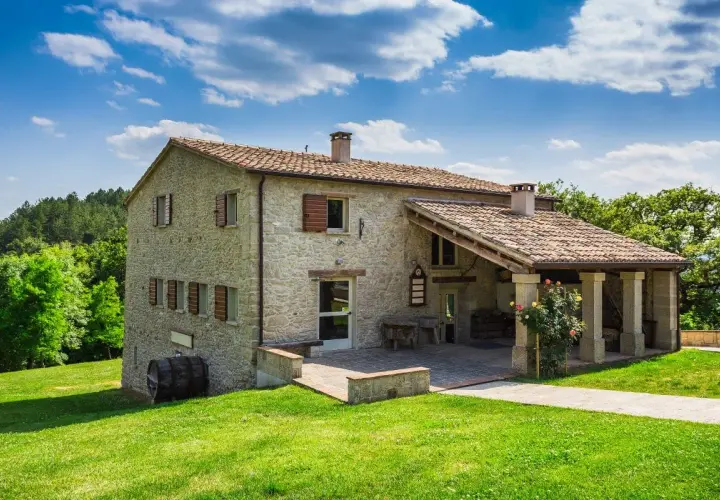 Belle ferme en pierre, rénovation réussie, avec un porche couvert et des roses épanouies. Un paysage verdoyant et un ciel bleu complètent ce tableau.