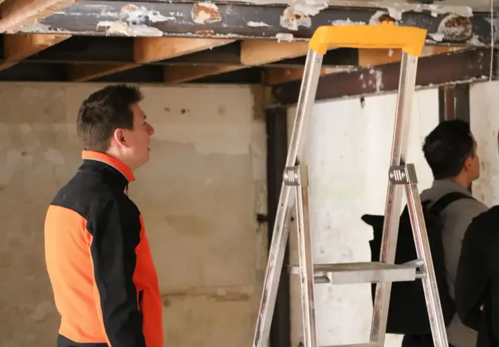 Un homme en veste orange et noire observe le plafond délabré d'un bâtiment, signe d'une future rénovation énergétique des bâtiments publics.