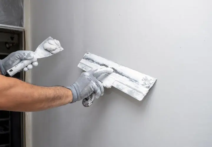 Mains gantées appliquant du machefer sur un mur gris avec des truelles.