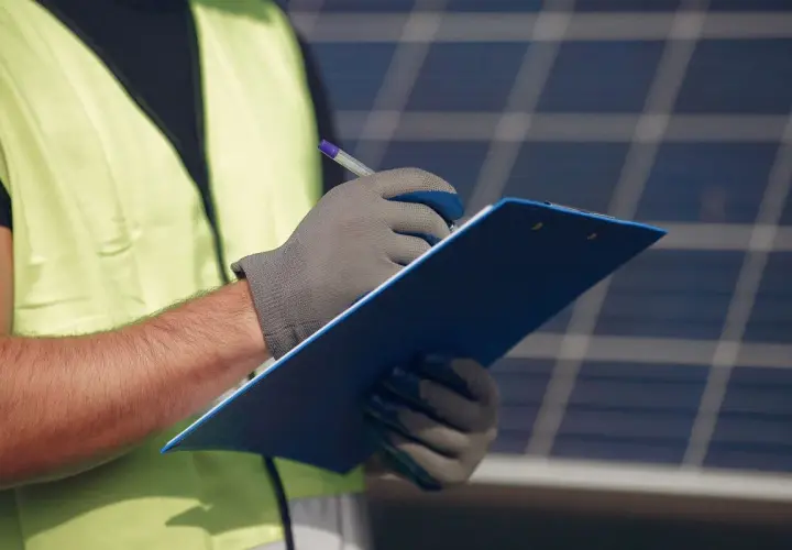 Un technicien en 50549, portant un gilet de sécurité, note des informations sur un presse-papiers bleu devant des panneaux solaires.