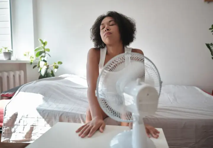 Une jeune femme en débardeur blanc, accablée par la chaleur de l'été, cherche un peu de fraîcheur devant un ventilateur. L'isolation de sa chambre ne suffit pas.