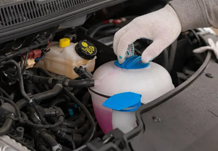 Main d'une personne portant un gant blanc ferme le bouchon d'un vase d'expansion rempli d'eau et de liquide de refroidissement rose.