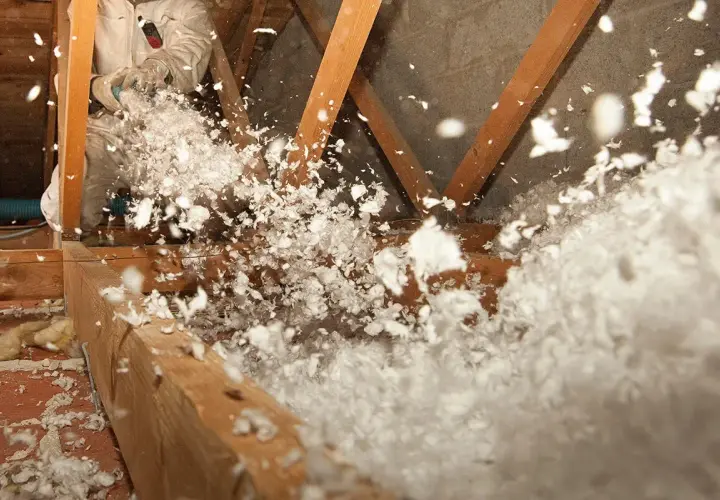 Un ouvrier en combinaison souffle de la laine de verre dans un grenier, créant un nuage blanc et cotonneux pour une isolation efficace.
