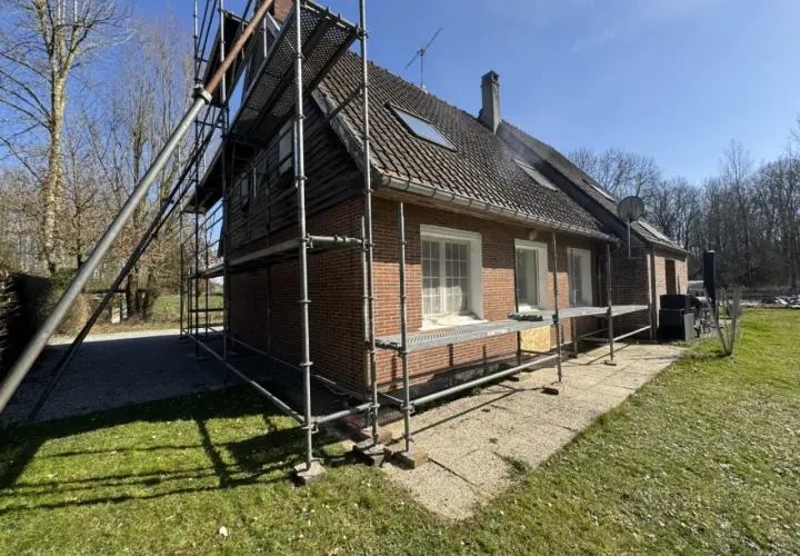 Échafaudage autour d'une maison en briques rouges, signe d'une rénovation maison de ville en cours. Le soleil brille sur le chantier.