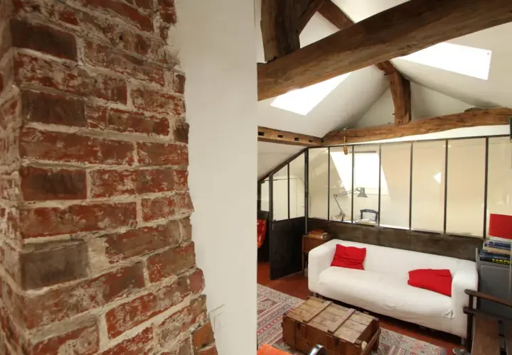 Murs en briques apparentes et poutres de bois, ambiance chaleureuse pour cette rénovation combles. Canapé blanc, coussins rouges, style loft.