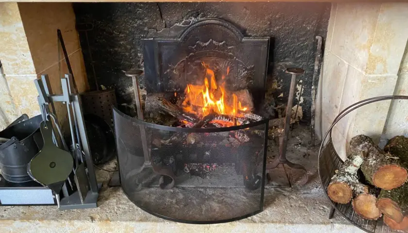 Un feu de bois crépite dans une cheminée. Des outils de cheminée et du bois sont à proximité, prêts à fabriquer un récupérateur de chaleur pour poêle à bois.