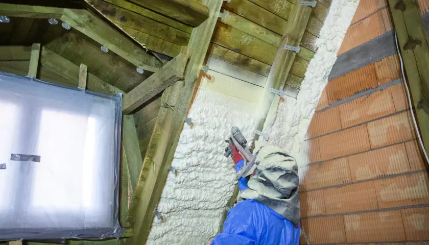 Un ouvrier en combinaison bleue applique du polyuréthane projeté sur la charpente d'un grenier, isolant ainsi les murs en briques et le toit.