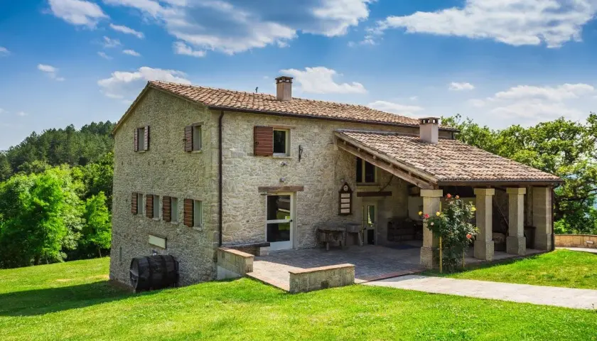Belle ferme en pierre, rénovation réussie, avec un porche couvert et des roses épanouies. Un paysage verdoyant et un ciel bleu complètent ce tableau.