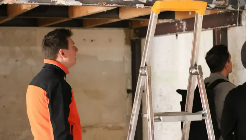 Un homme en veste orange et noire observe le plafond délabré d'un bâtiment, signe d'une future rénovation énergétique des bâtiments publics.
