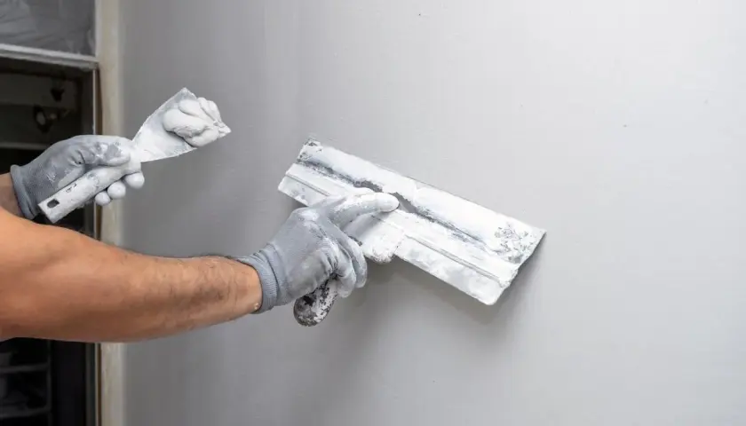 Mains gantées appliquant du machefer sur un mur gris avec des truelles.
