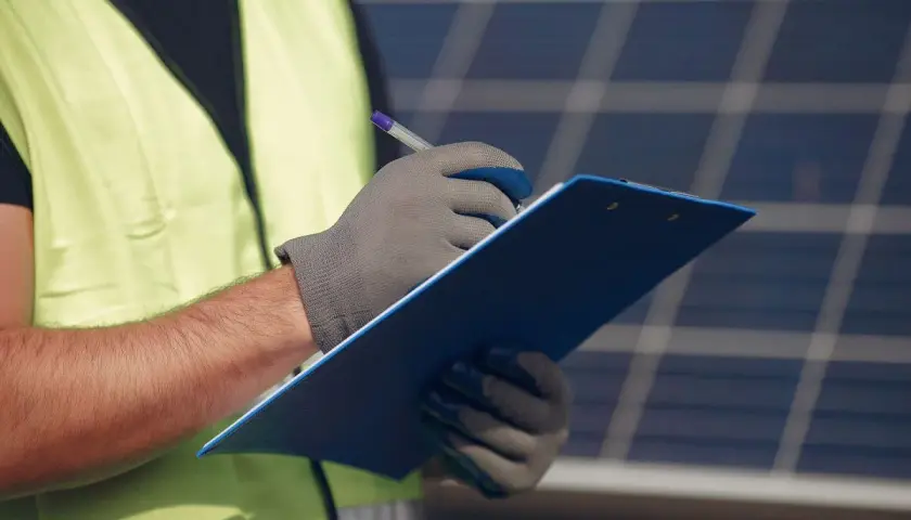 Un technicien en 50549, portant un gilet de sécurité, note des informations sur un presse-papiers bleu devant des panneaux solaires.