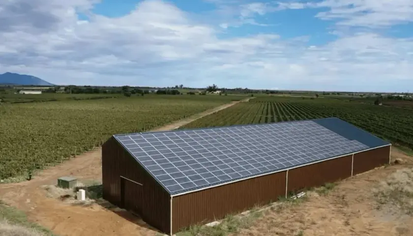 Vue aérienne d'un hangar brun avec un toit couvert de panneaux photovoltaïques, entouré de vignobles.