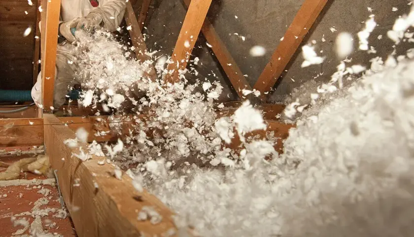 Un ouvrier en combinaison souffle de la laine de verre dans un grenier, créant un nuage blanc et cotonneux pour une isolation efficace.