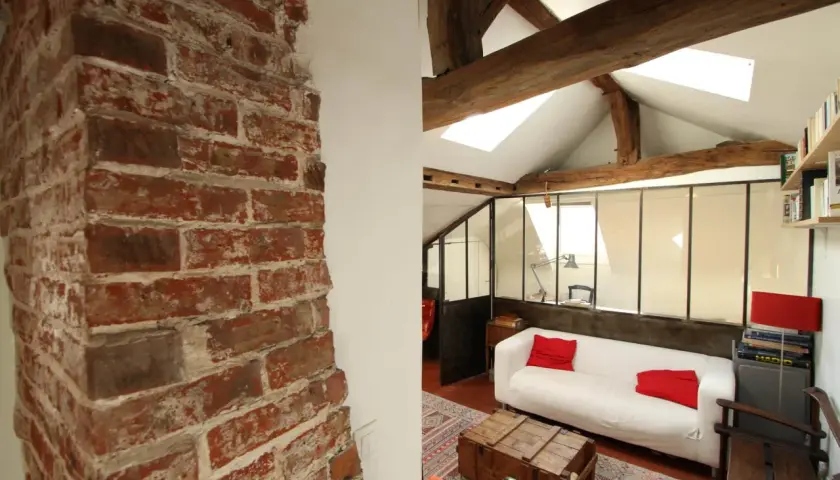 Murs en briques apparentes et poutres de bois, ambiance chaleureuse pour cette rénovation combles. Canapé blanc, coussins rouges, style loft.