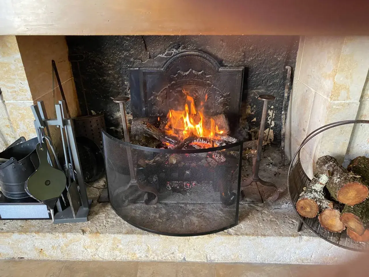 Un feu de bois crépite dans une cheminée. Des outils de cheminée et du bois sont à proximité, prêts à fabriquer un récupérateur de chaleur pour poêle à bois.