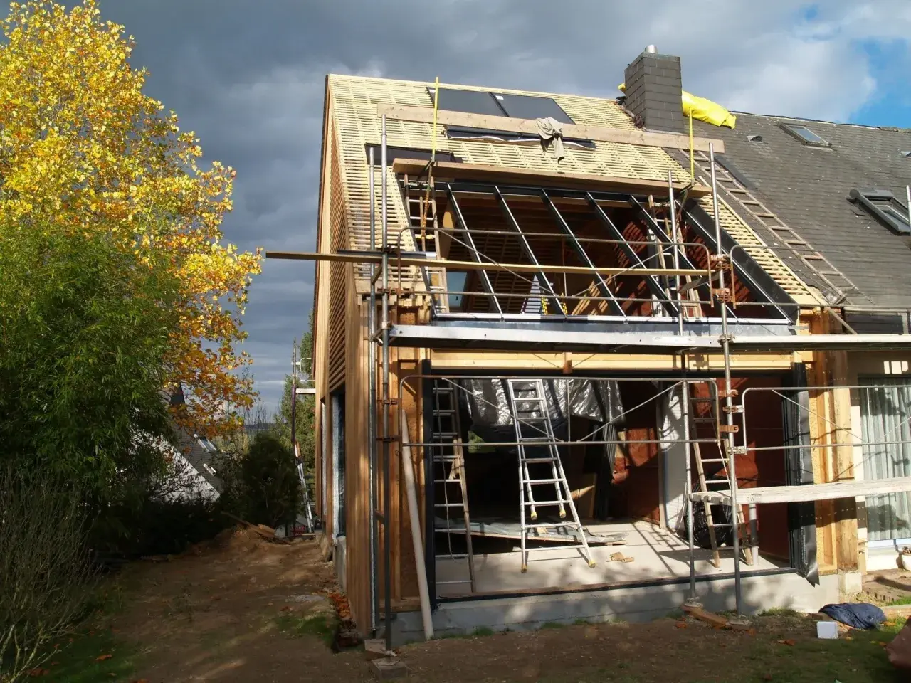 Chantier de rénovation maison : échafaudages, charpente apparente, et une partie de la façade en bois. Le ciel est menaçant.