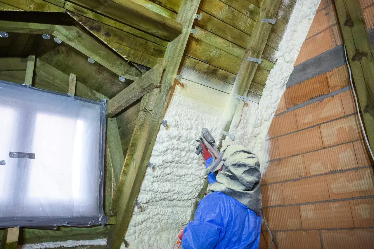 Un ouvrier en combinaison bleue applique du polyuréthane projeté sur la charpente d'un grenier, isolant ainsi les murs en briques et le toit.