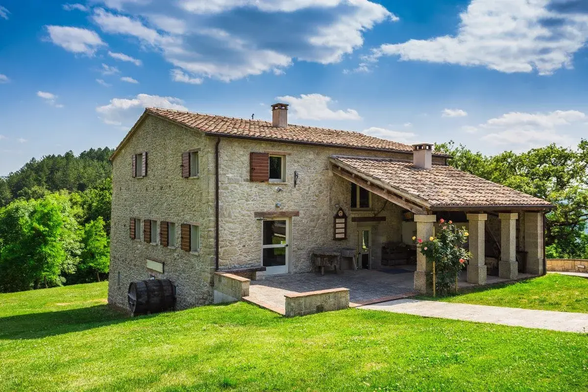 Belle ferme en pierre, rénovation réussie, avec un porche couvert et des roses épanouies. Un paysage verdoyant et un ciel bleu complètent ce tableau.
