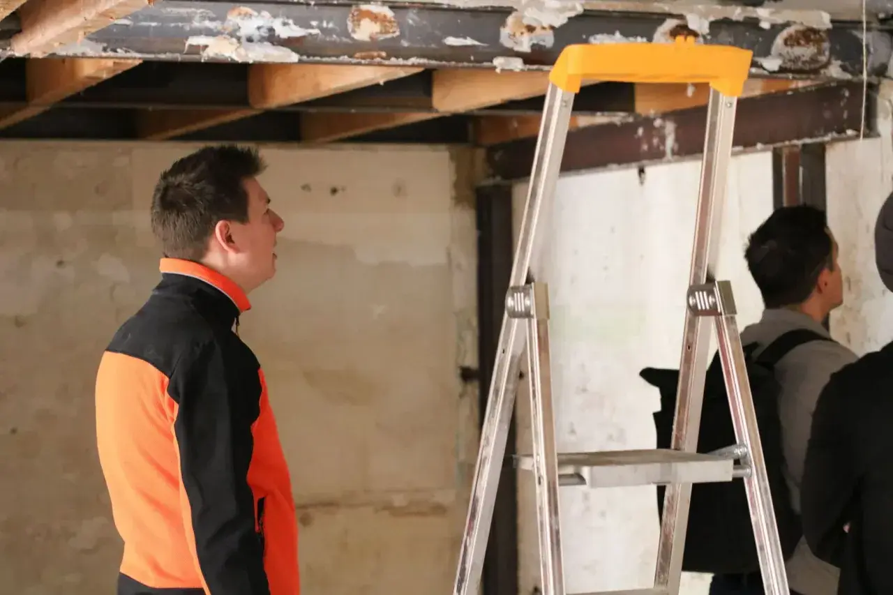 Un homme en veste orange et noire observe le plafond délabré d'un bâtiment, signe d'une future rénovation énergétique des bâtiments publics.