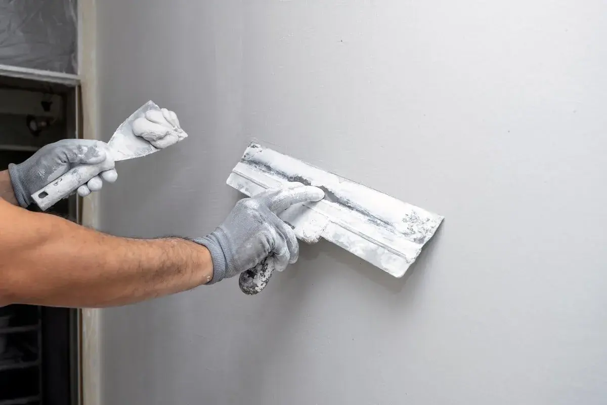 Mains gantées appliquant du machefer sur un mur gris avec des truelles.