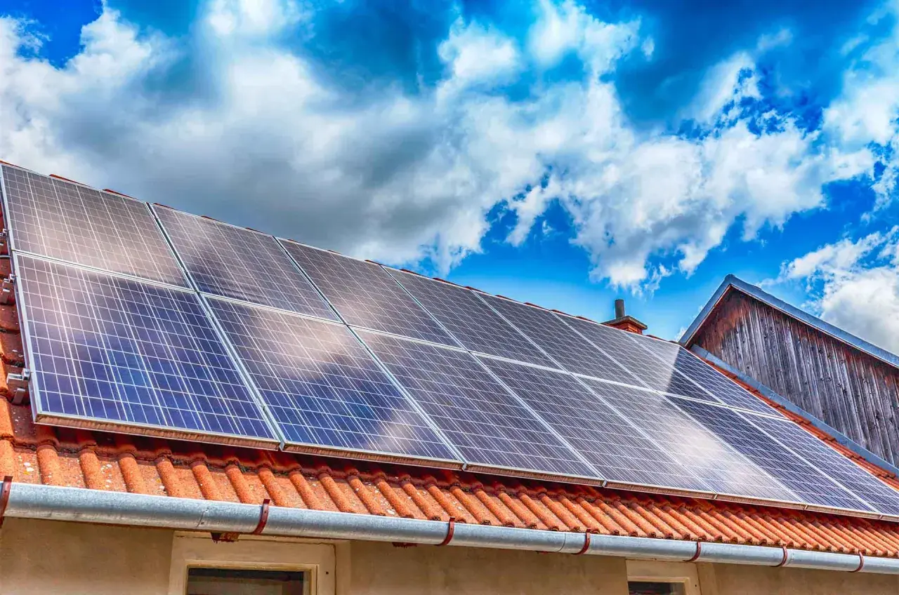 Panneaux solaires sur un toit de tuiles rouges sous un ciel bleu et nuageux. Une installation qui peut influencer le coût de l'assurance photovoltaïque.