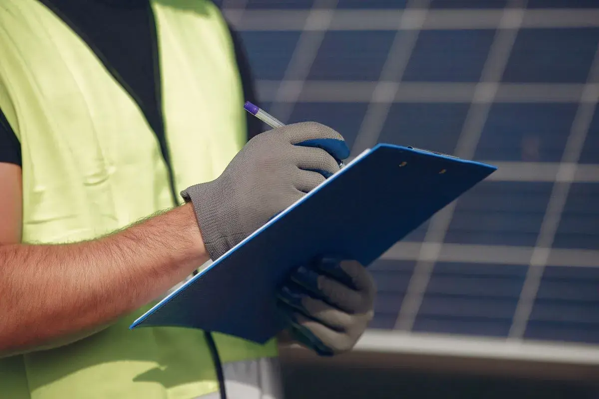 Un technicien en 50549, portant un gilet de sécurité, note des informations sur un presse-papiers bleu devant des panneaux solaires.