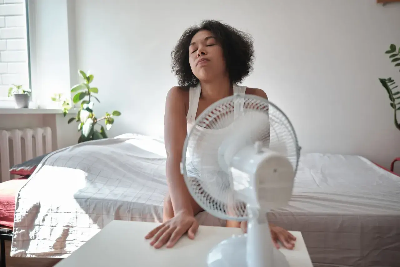 Une jeune femme en débardeur blanc, accablée par la chaleur de l'été, cherche un peu de fraîcheur devant un ventilateur. L'isolation de sa chambre ne suffit pas.