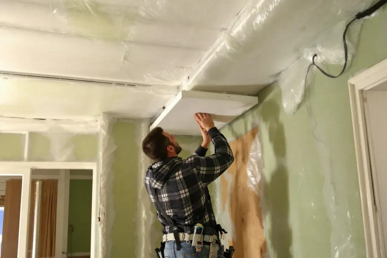 Un homme installe un panneau au plafond, entouré de bâches. Attention, la laine de verre peut présenter un danger pour les poumons si mal manipulée.