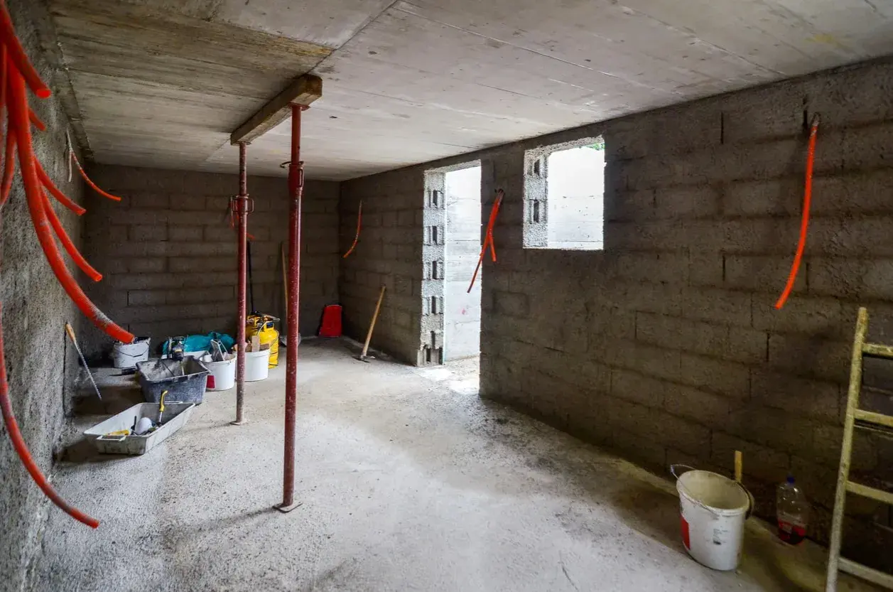 Intérieur brut d'une cave en construction, avec des tuyaux orange, des seaux et des étais. L'isolation de cette cave est en cours.