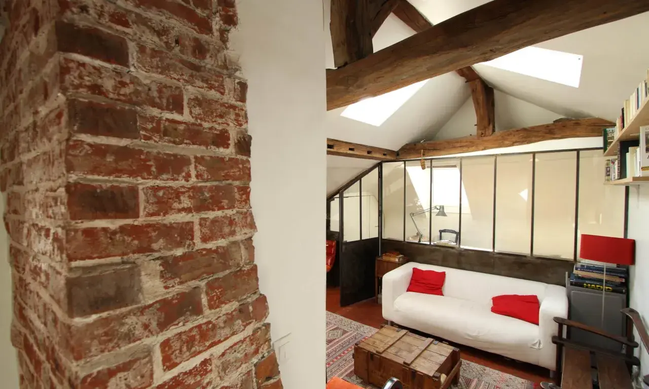 Murs en briques apparentes et poutres de bois, ambiance chaleureuse pour cette rénovation combles. Canapé blanc, coussins rouges, style loft.