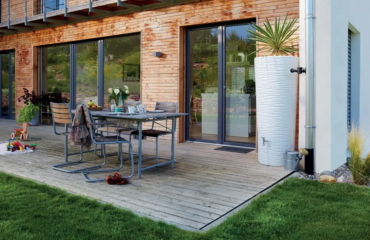 Installation récupérateur eau de pluie moderne, avec une cuve blanche texturée et un robinet, à côté d'une gouttière sur une maison en bois.