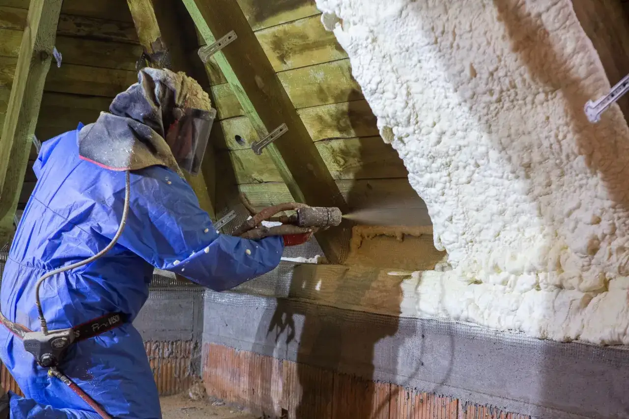 Un technicien en combinaison bleue applique du polyuréthane projeté sur les combles pour une isolation efficace.