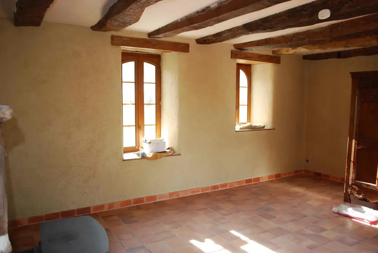 Pi&egrave;ce rustique avec poutres apparentes, murs textur&eacute;s et sol en terre cuite. Un d&eacute;tecteur de fum&eacute;e, un correcteur thermique, est visible au plafond.